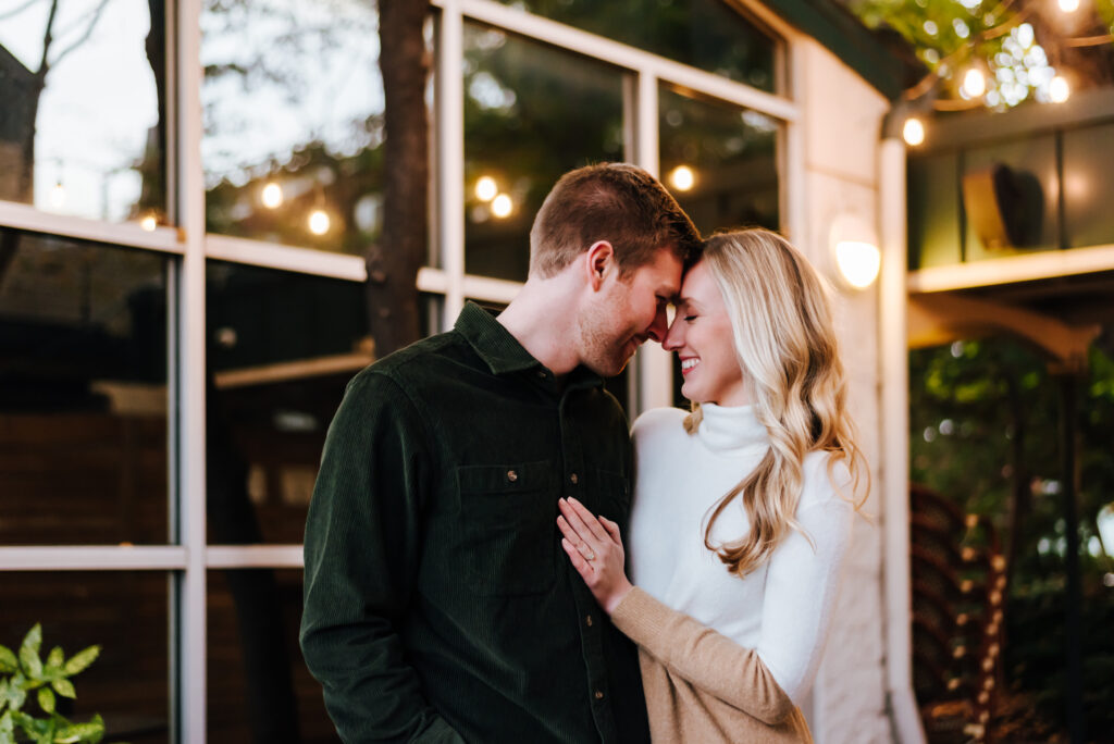 Couple embraces in front of a glass building with string lights