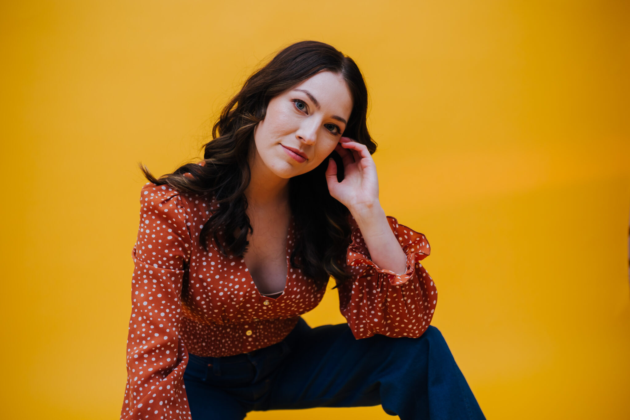 Johanna, wearing a 1970s inspired ruffle top and wide leg jeans, poses with her arm resting on her knee and tilts her head sideways with a smile