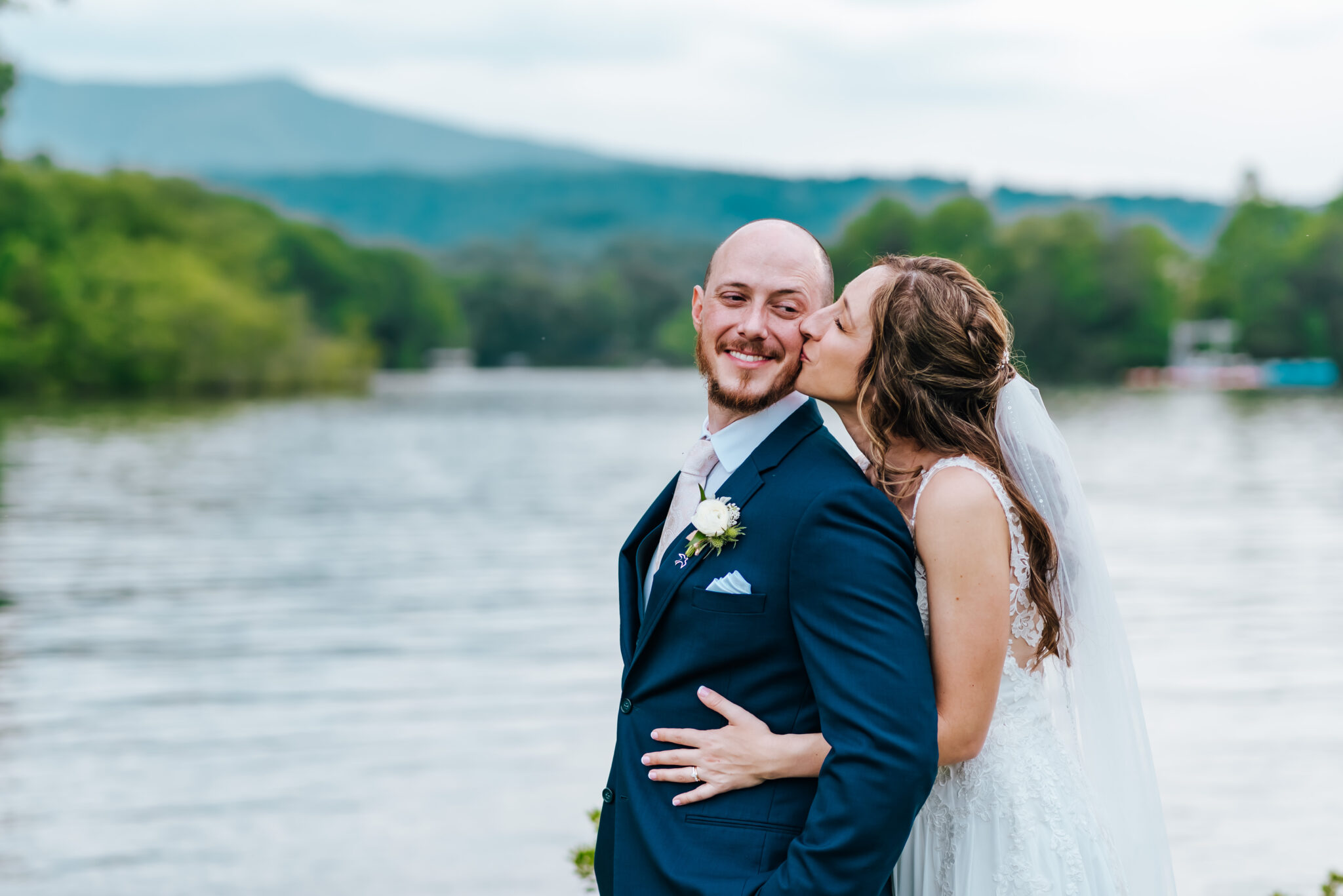 Bride kisses groom from behind as they stand in front of a mountain lake