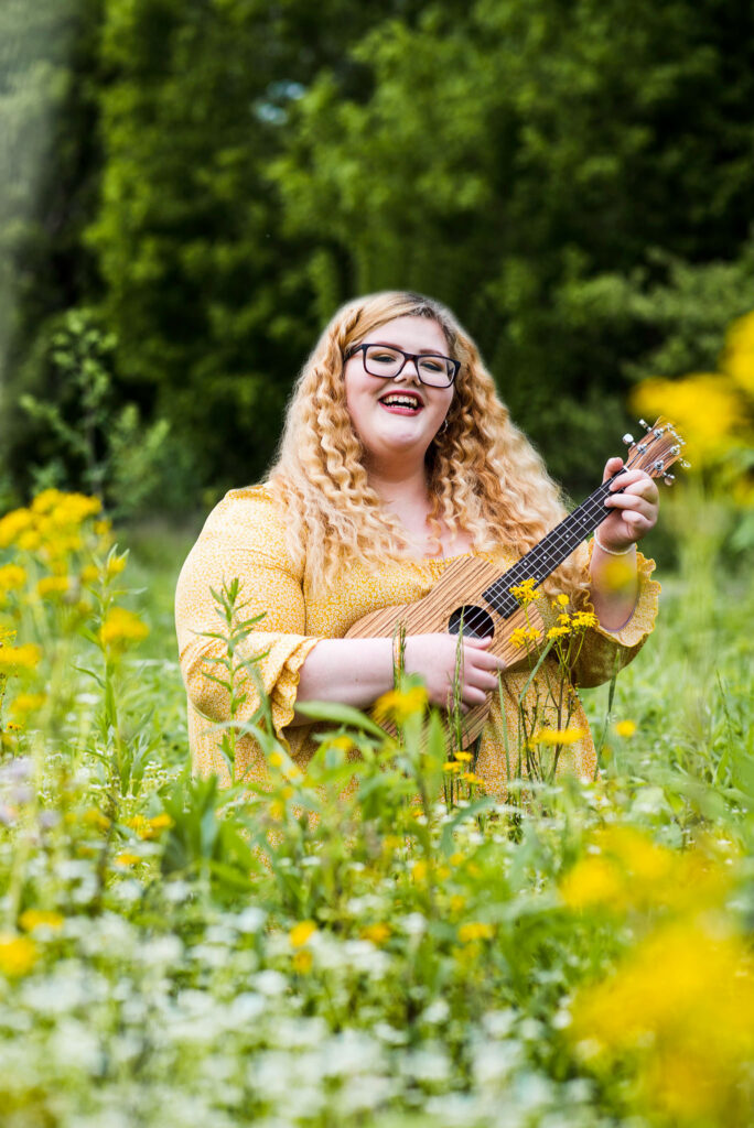 High School girl in a yellow dress smiles and plays the Ukulele in a flower field