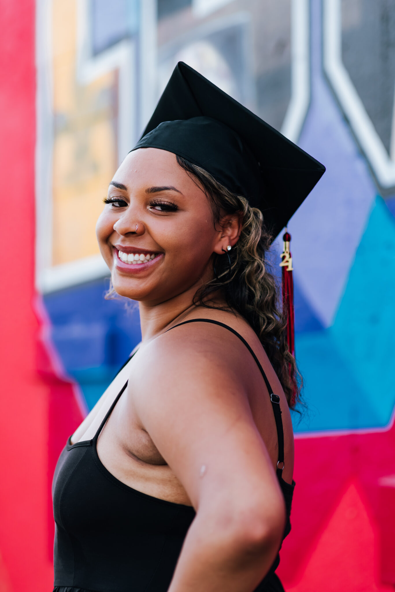 Teen in a black dress and graduation cap smiles in front of a colorful mural