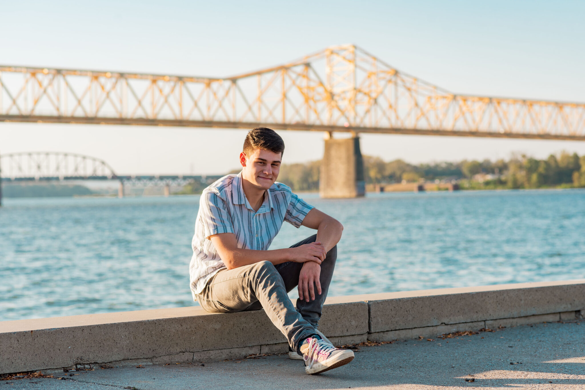 Teen boy sits in front of the Ohio River with yellow bridge behind him at sunset