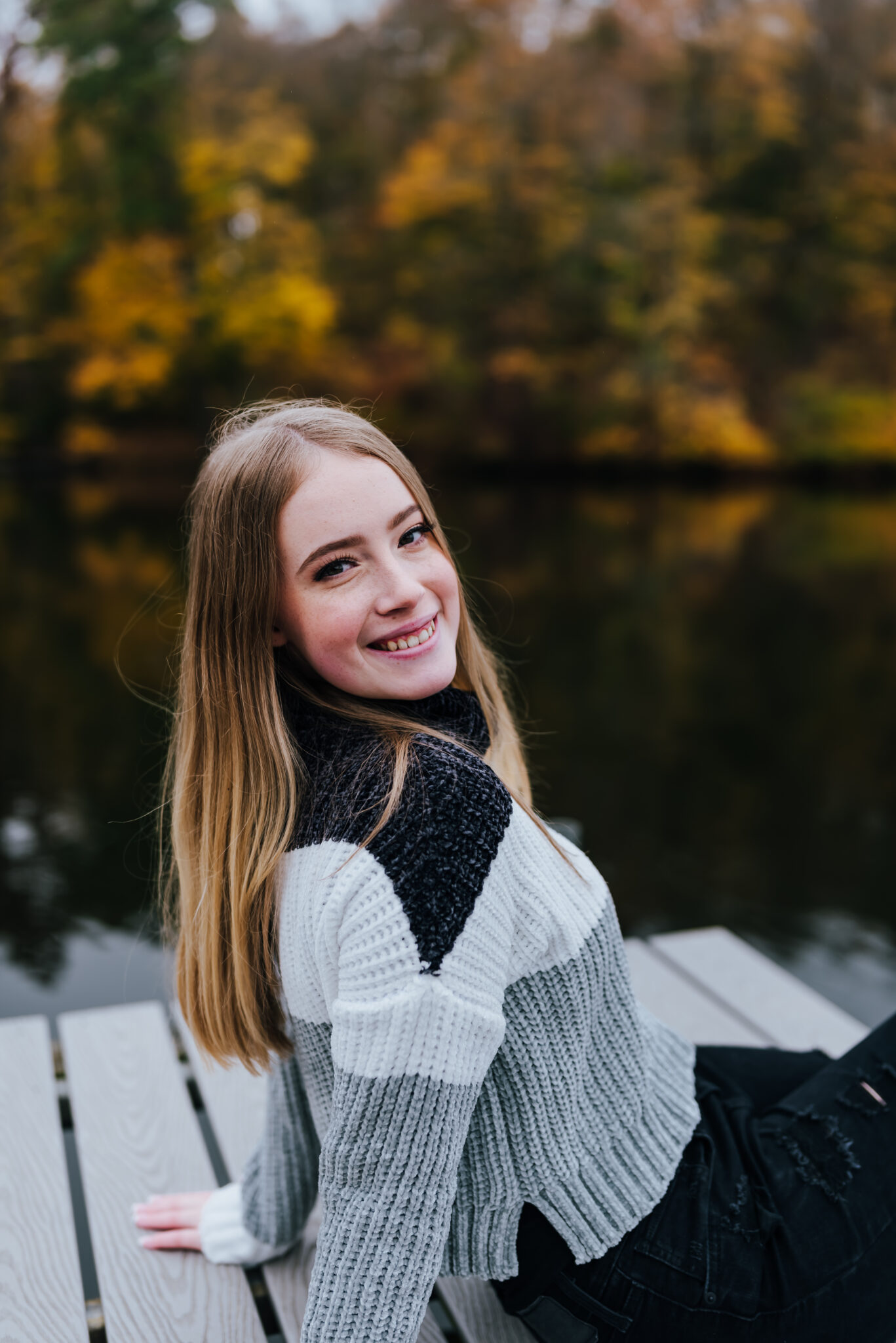 Girl in sweater smiles and sits on dock with tall leaves behind her