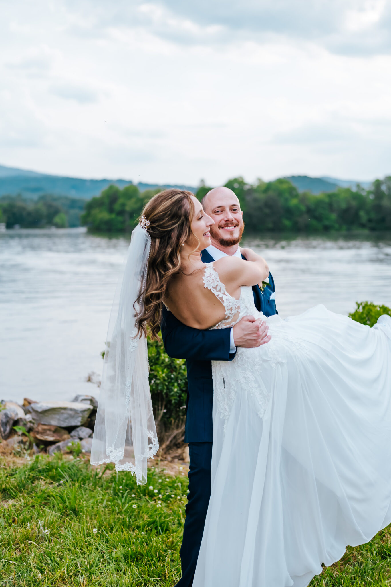 A groom carries his bride in her wedding dress and veil as they walk in front of a beautiful mountain lake