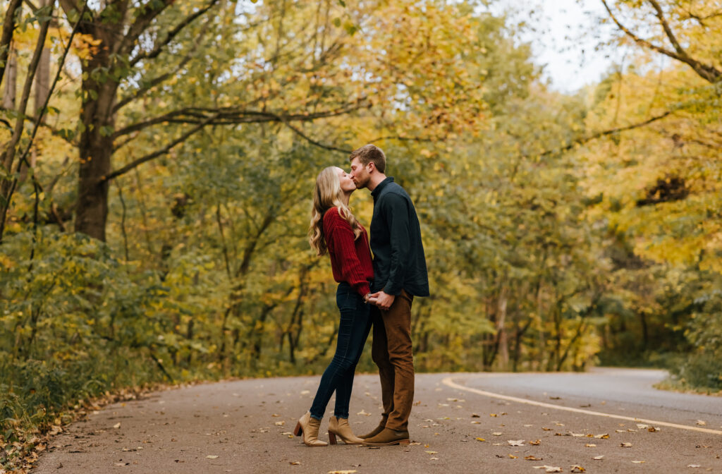 Couple kisses on an empty road surrounded by fall trees with yellow leaves