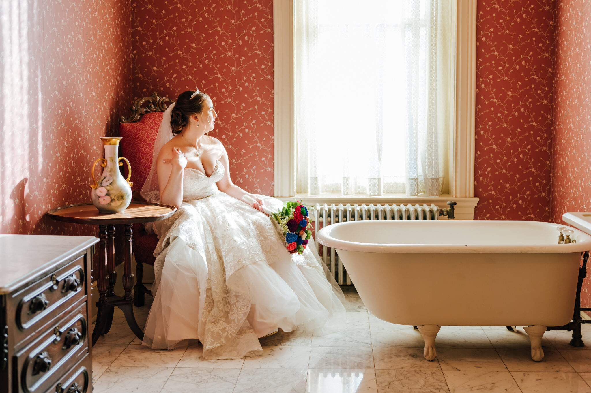 A bride sits in an elegant chair in a historic bathroom with pink wallpaper. She gazes out the window.