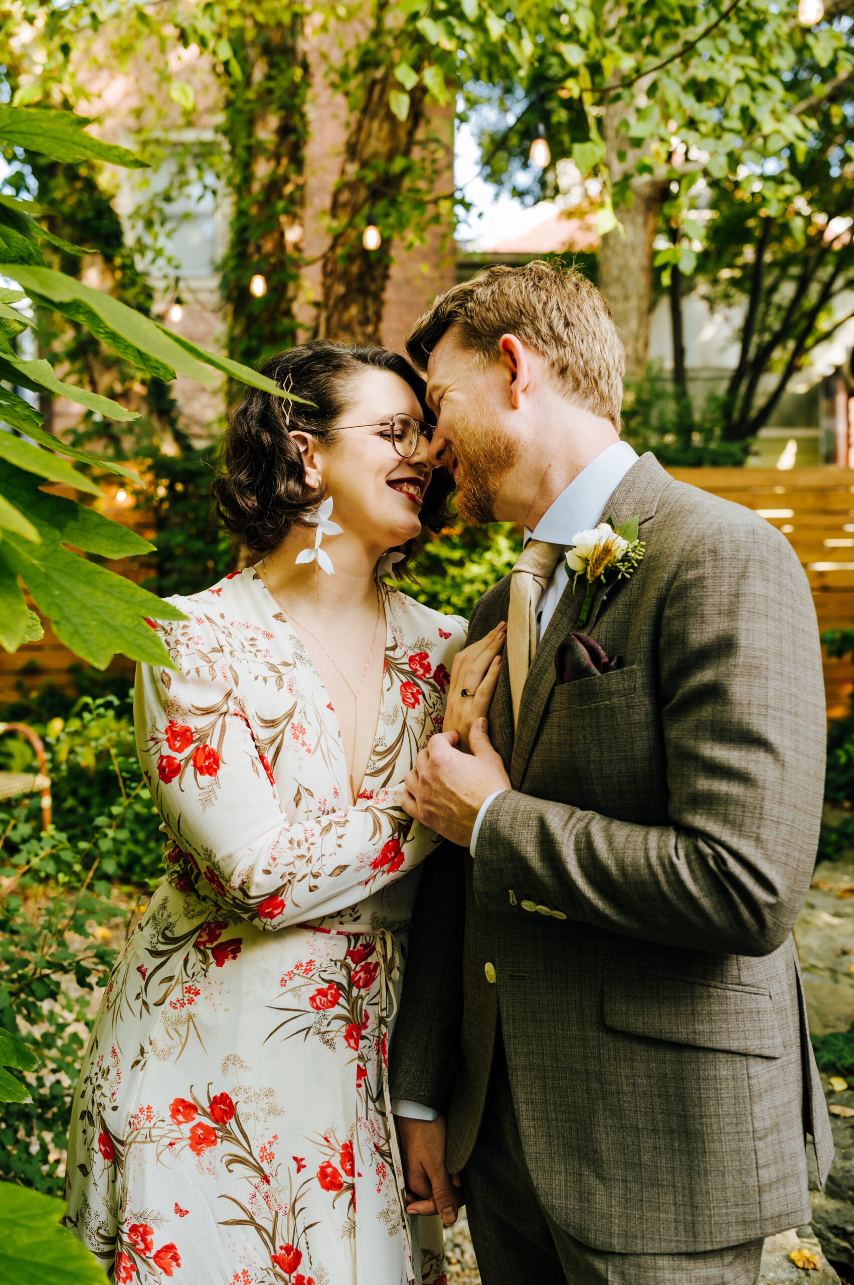 Couple embraces and smiles at one another. They are standing in a lush garden with wedding attire.