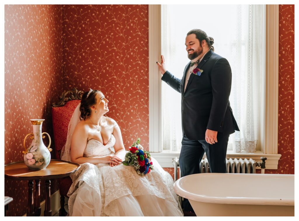 A couple in wedding attire looks at each other as they sit in a historic wallpapered room