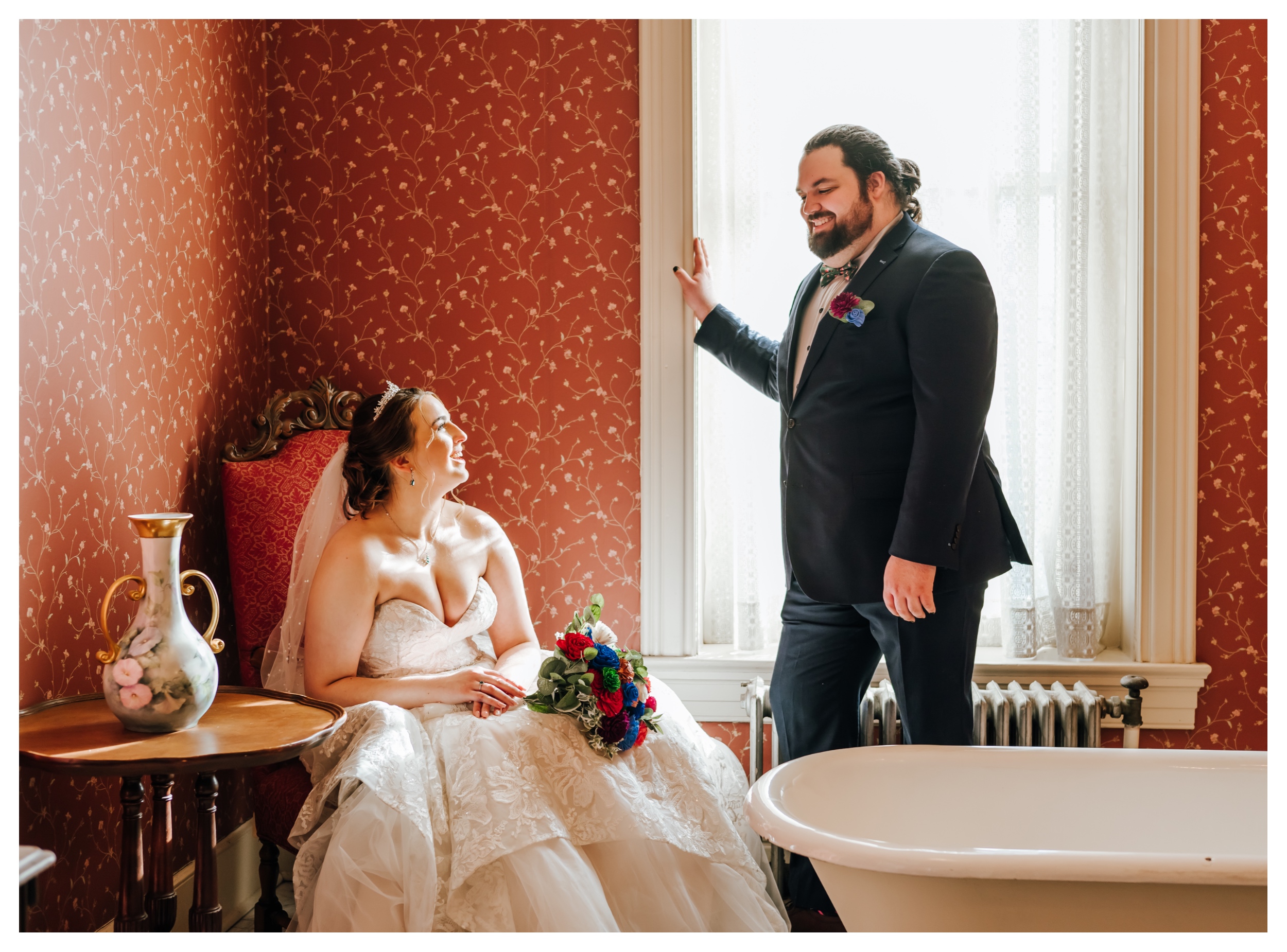 A couple in wedding attire shares a quiet moment in a historic wallpapered room with natural light leaking in