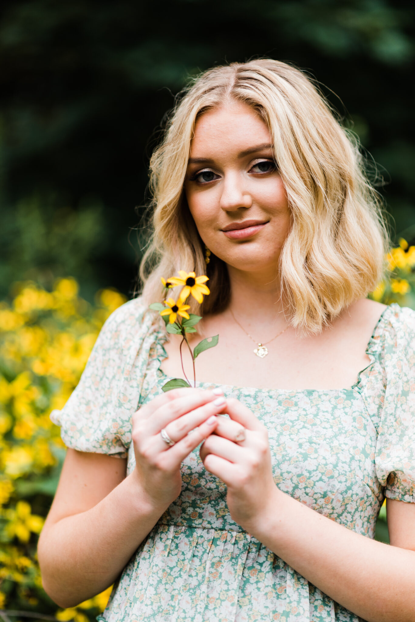 A girl with short blond hair and a green dress holds a few yellow flowers in her hands