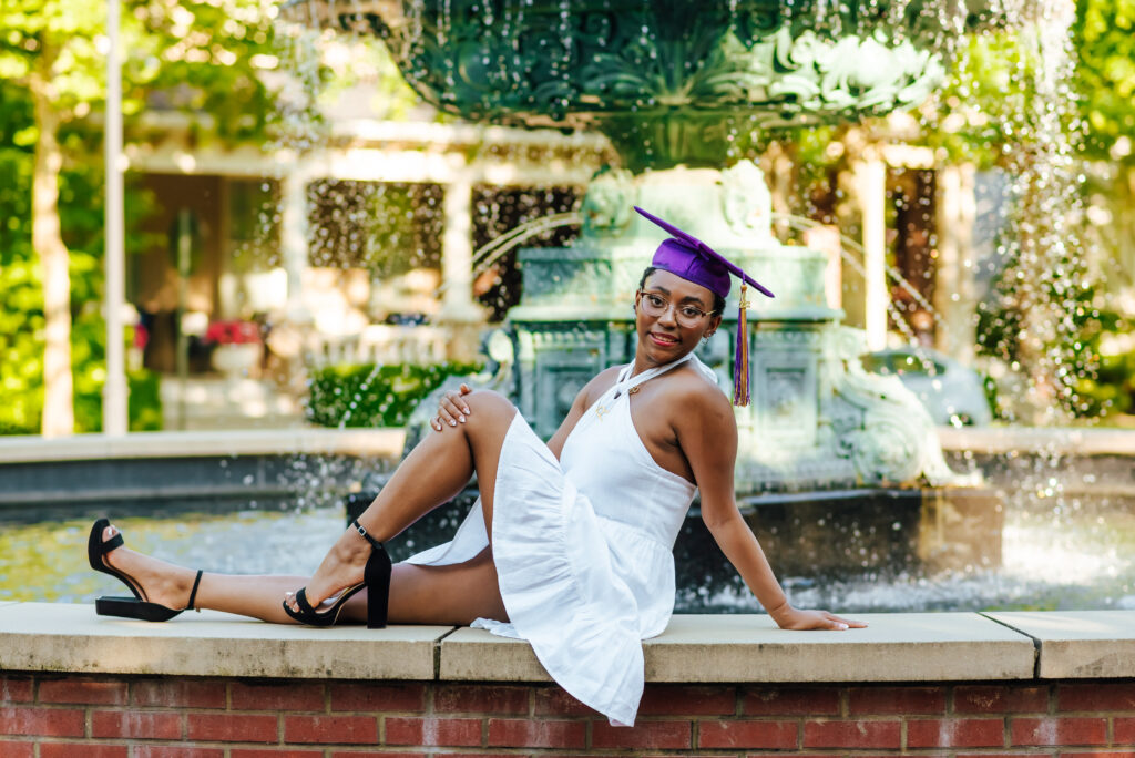 A graduate of Male High School wearing a white dress and her purple graduation cap, sits in front of a green fountain outdoors