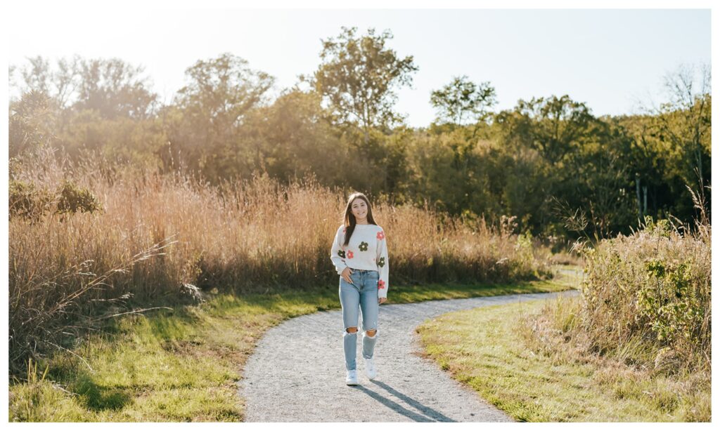 A girl in a flower sweater and jeans walks through a tall grassy park field