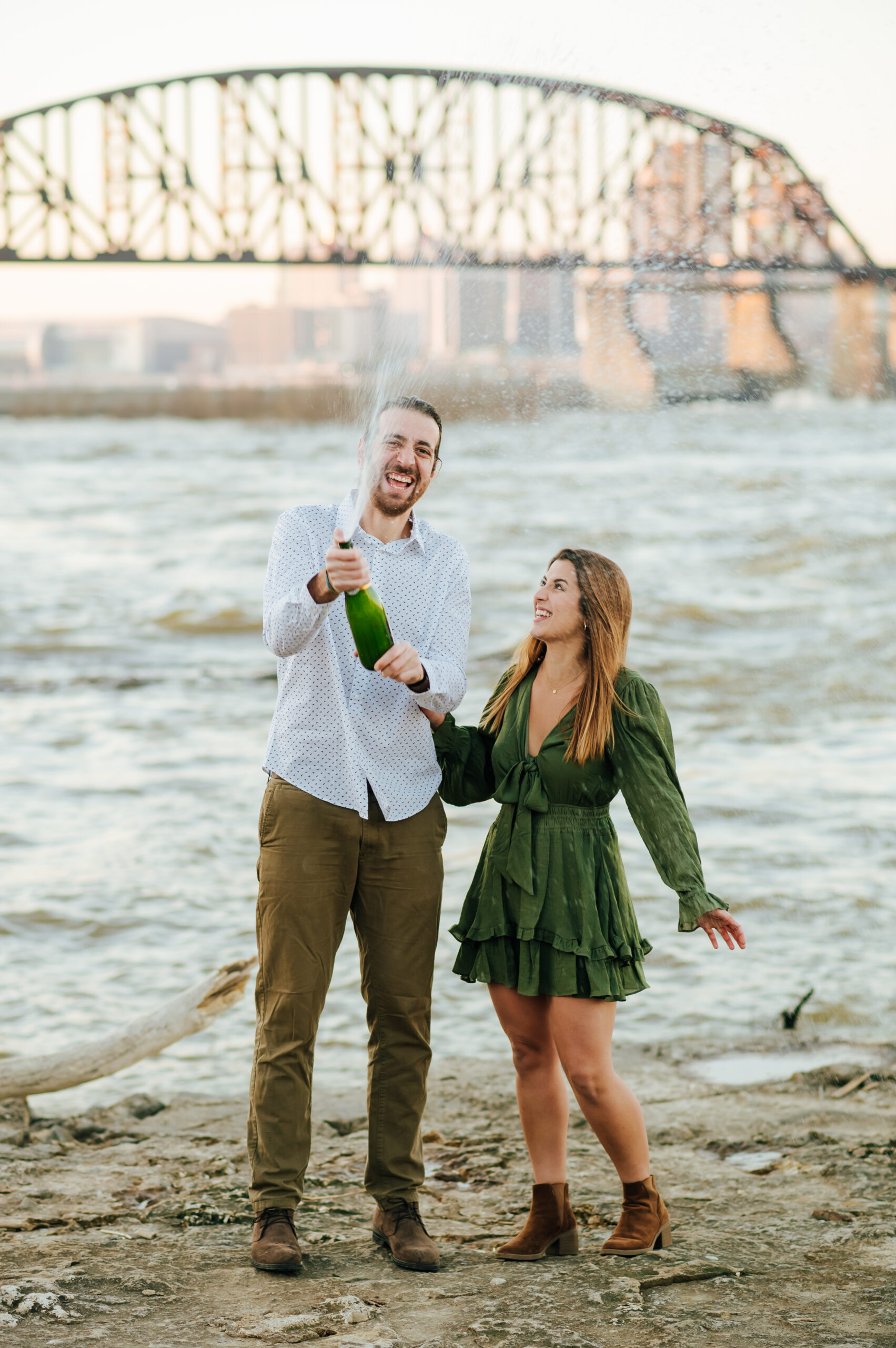 A couple sprays champagne in front of a beautiful bridge with water surrounding them