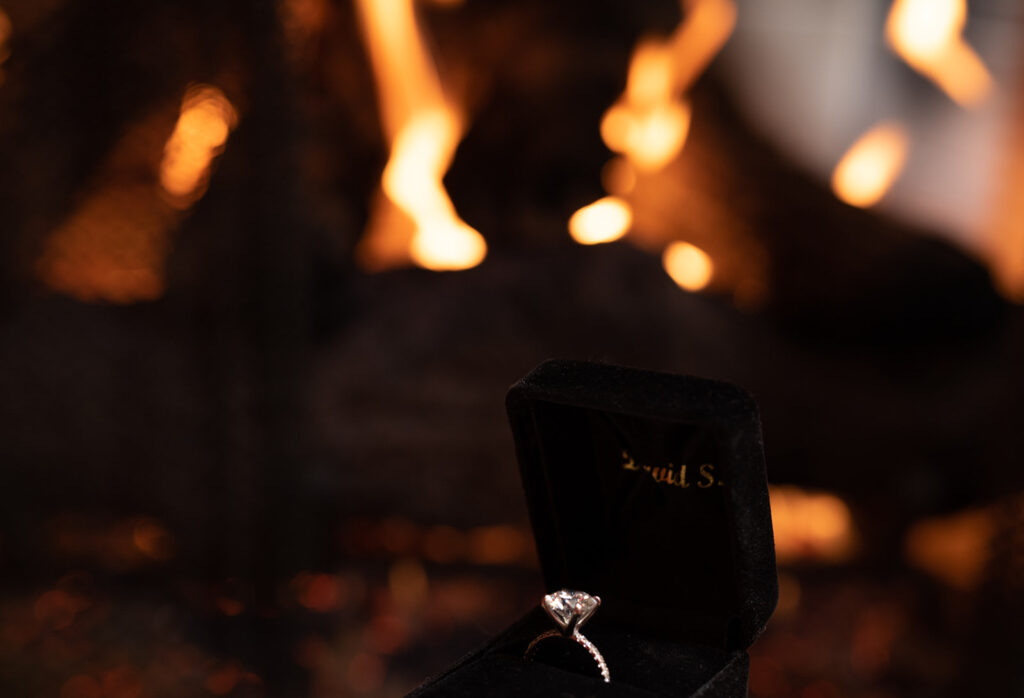 A beautiful black ring box with a solitaire diamond rests on the fireplace with a warm fire going