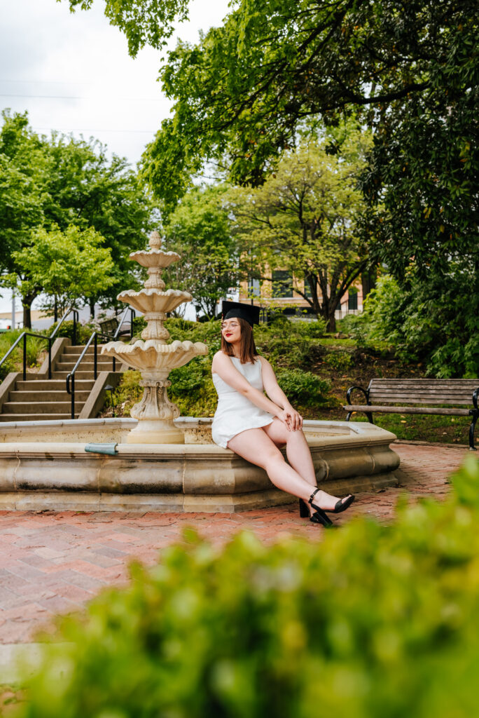 A girl in a white dress with a black graduation cap sits on the edge of a historic fountain surrounded by greenery