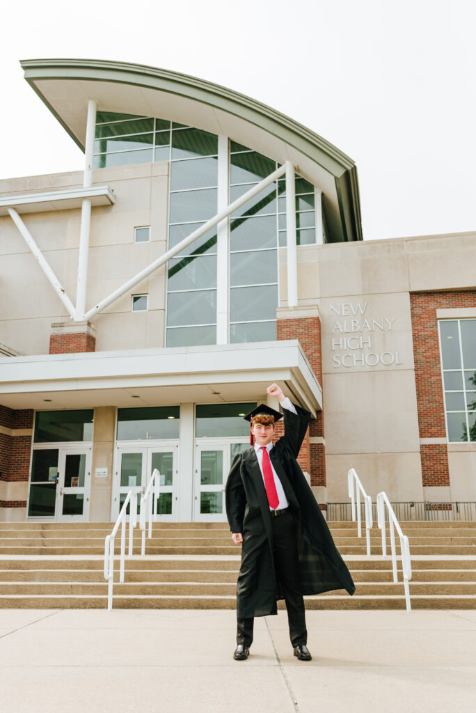 A high school grad does the classic Breakfast Club pose with his fist in the air wearing a cap and gown