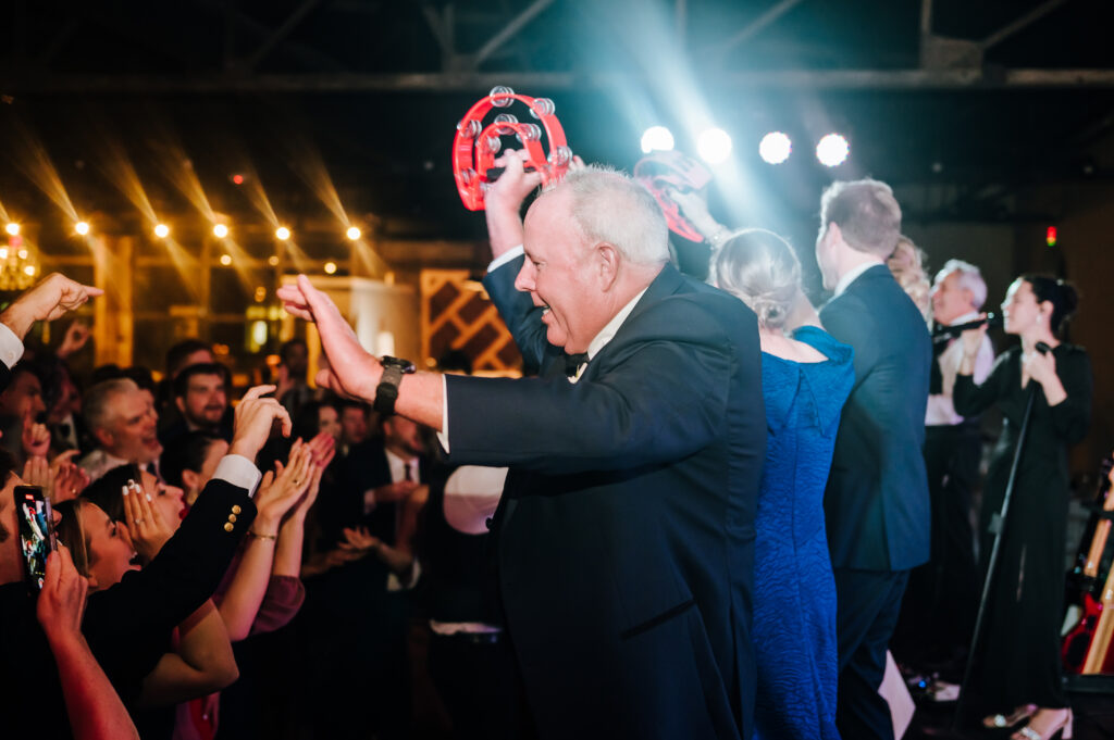 The groom's father shakes a red tambourine on stage at his son's wedding