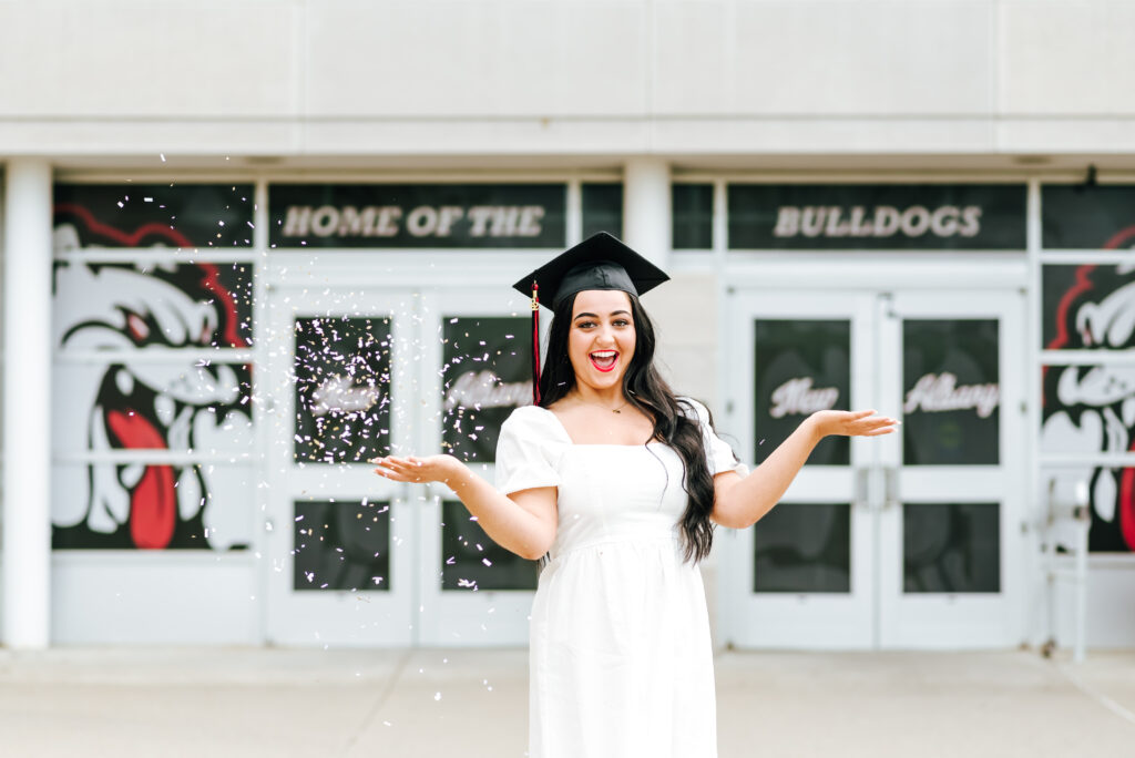 An excited high school grad in a white dress throws confetti in the air to celebrate