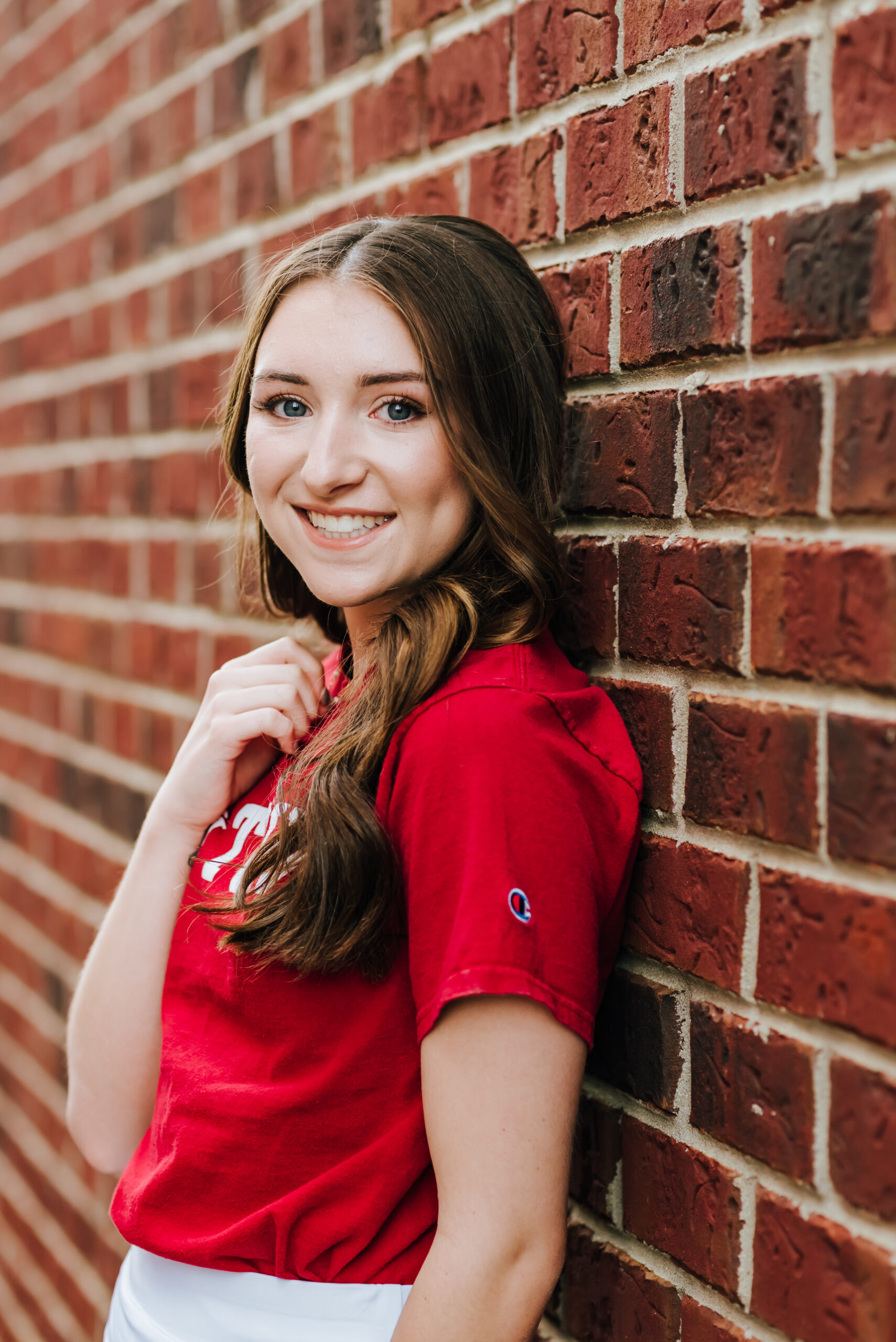 A high school senior in a red shirt smiles while leaning against a brick wall