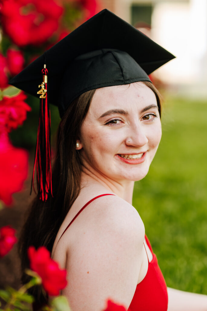 A high school graduate smiles wearing a red jumpsuit and a black graduation cap. She sits surrounded by a field of pink flowers