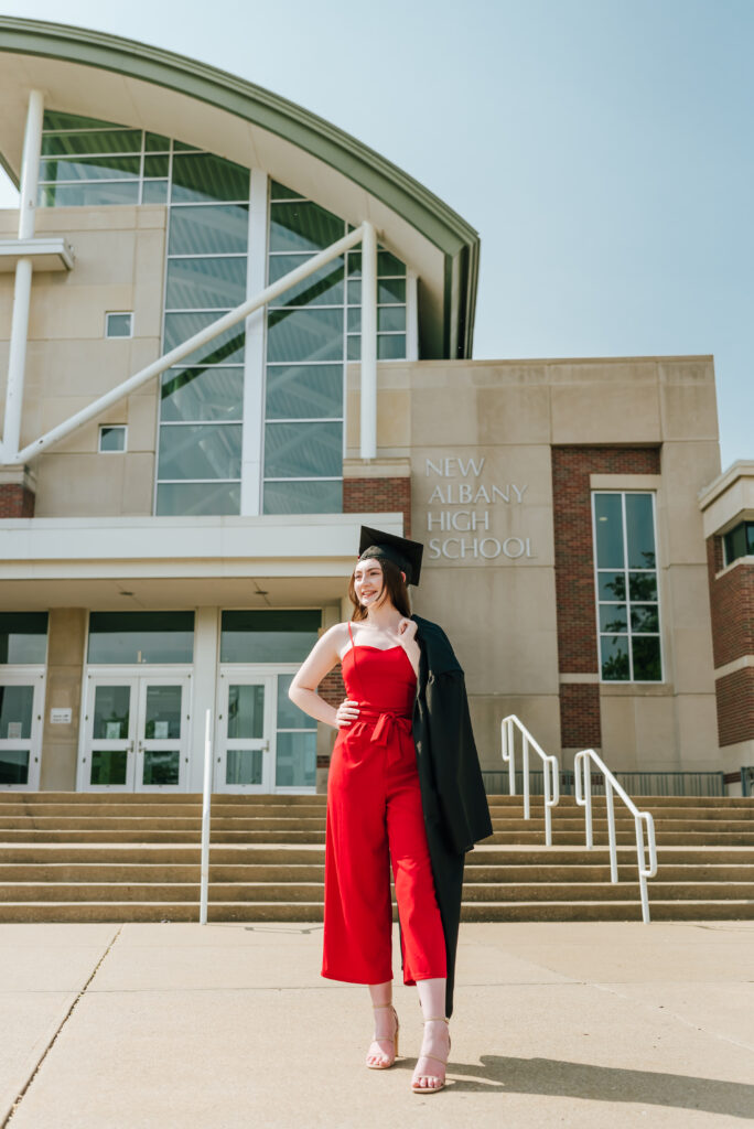 A high school graduate in a red jumpsuit poses confidently with her cap and gown in front of her school