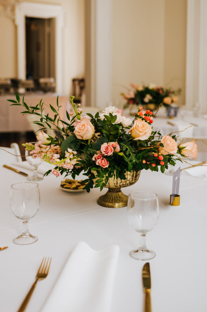 A peach toned bouquet sits in a golden vase on a decorated wedding reception table