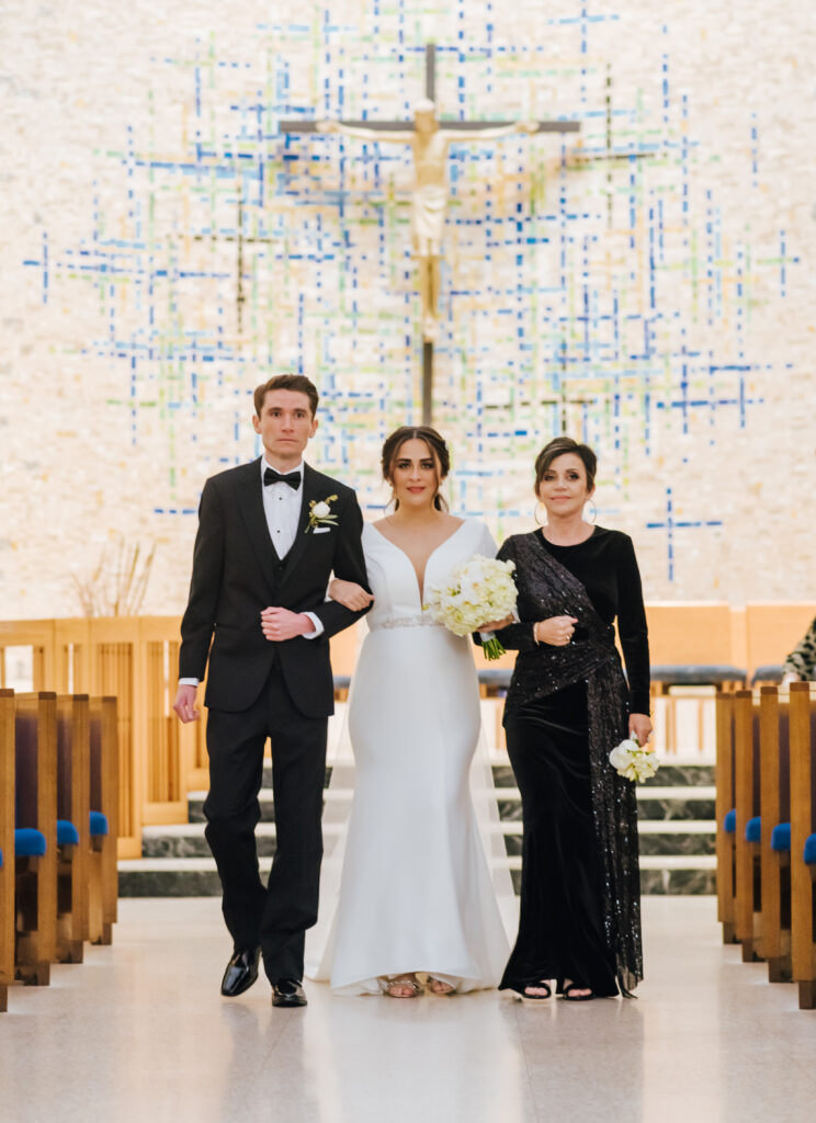Bride walks down the aisle, accompanied by her brother and mother