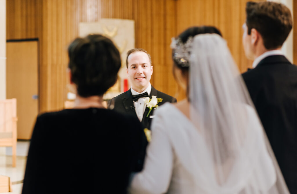 A guest's perspective photo of the groom in focus as he smiles at his bride