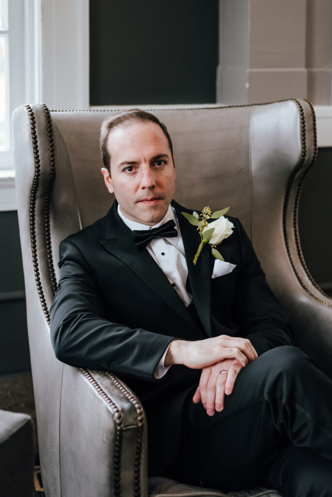 The stylish groom sits in a fancy armchair in his black tux