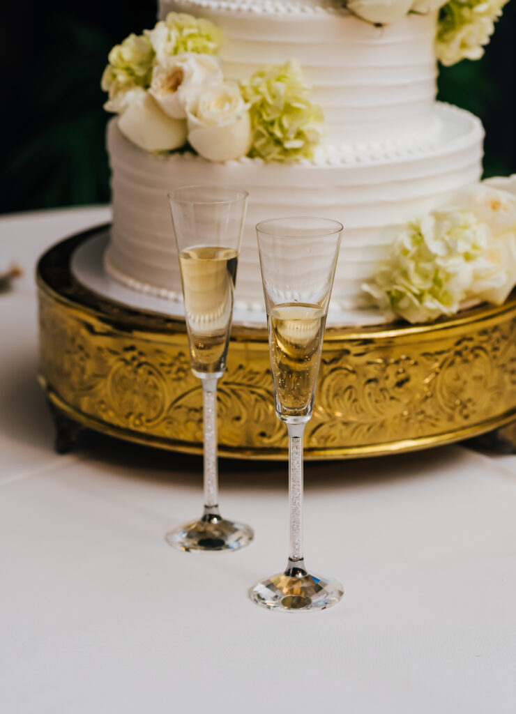 A close up detail photo of the two vintage champagne glasses in front of their white cake on a golden cake stand