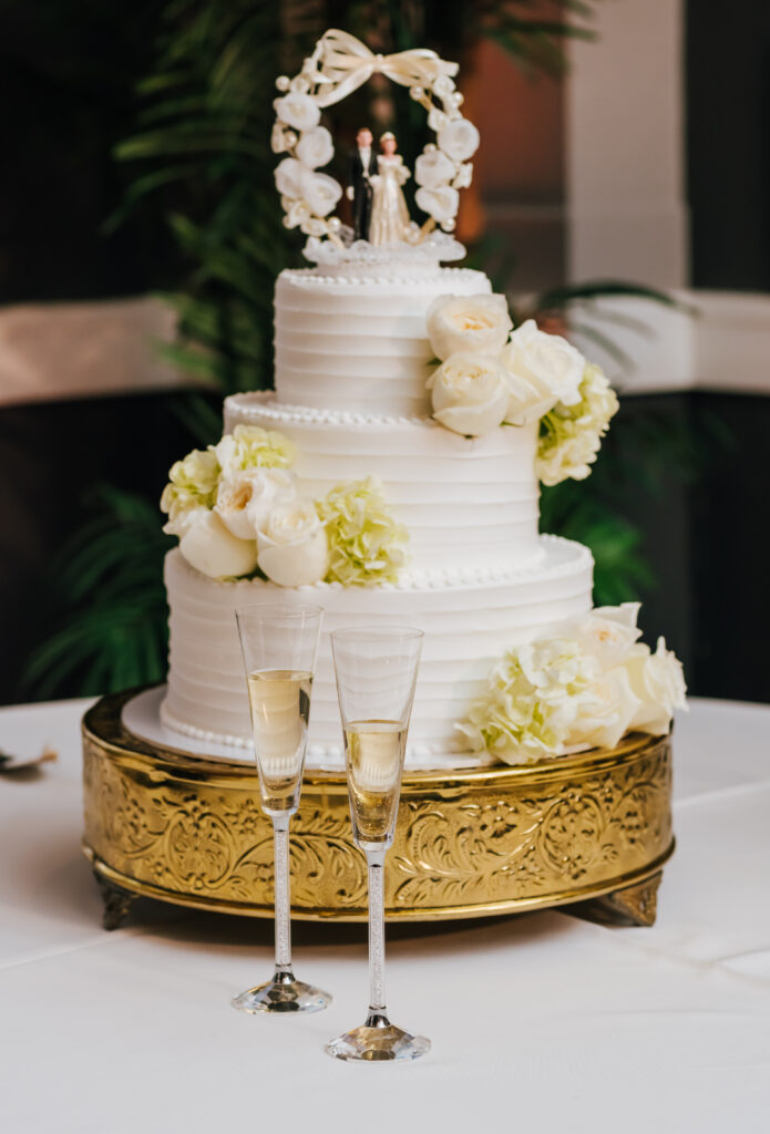 A photo of the couple's three tier white cake with flowers and a cake topper from the groom's grandparents. In front of the cake are two champagne glasses.