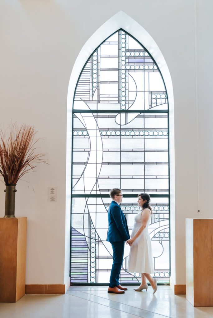 The newlyweds face each other and hold hands in front of tall stained glass windwos