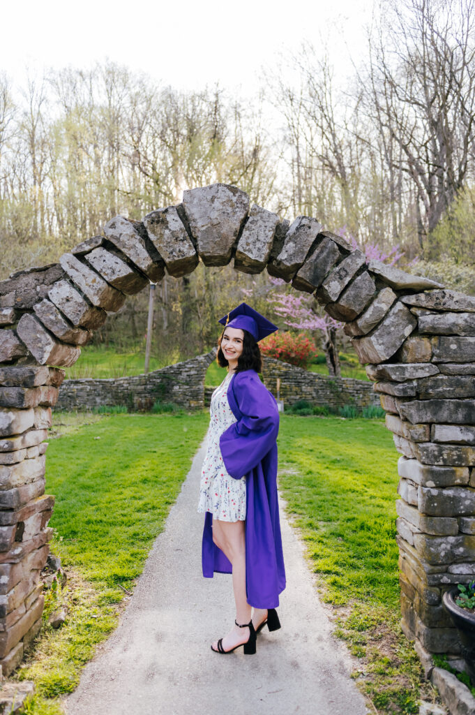 A graduate poses with her purple cap and gown under a stone archway