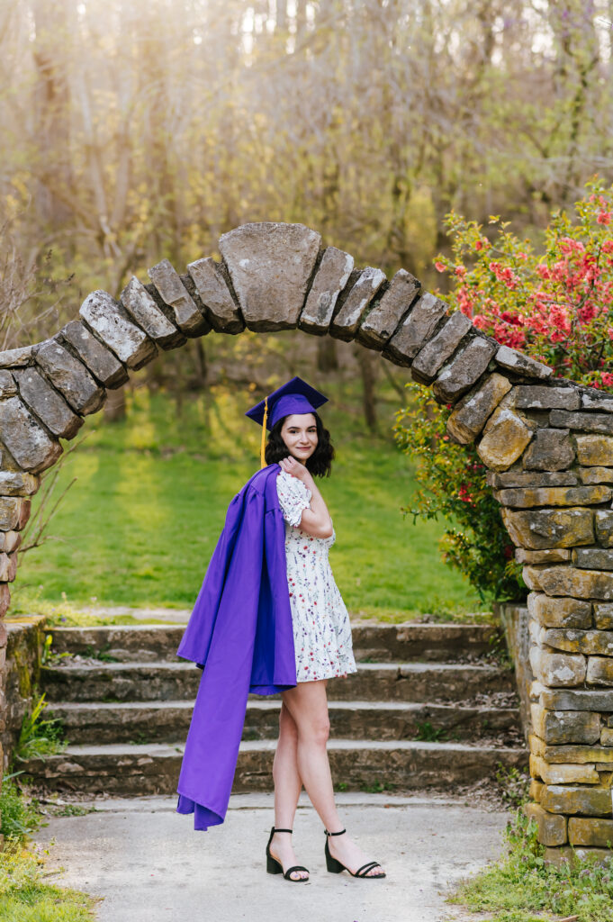 A senior wearing a white floral dress drapes her purple graduation gown over her shoulder and wears her cap.