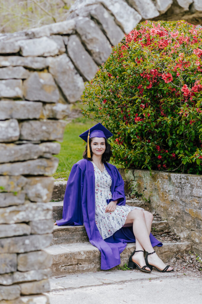 A graduate sits on a staircase under a stone arch wearing her purple cap and gown