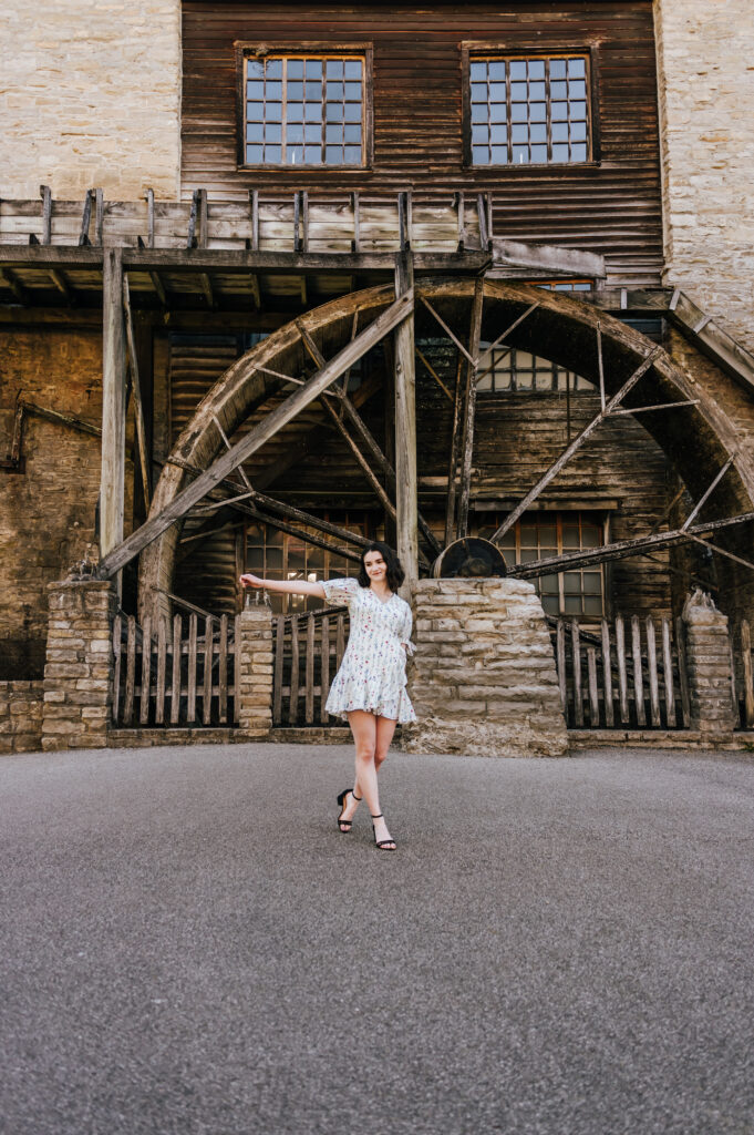A senior in a white floral dress poses in front of an old stone mill