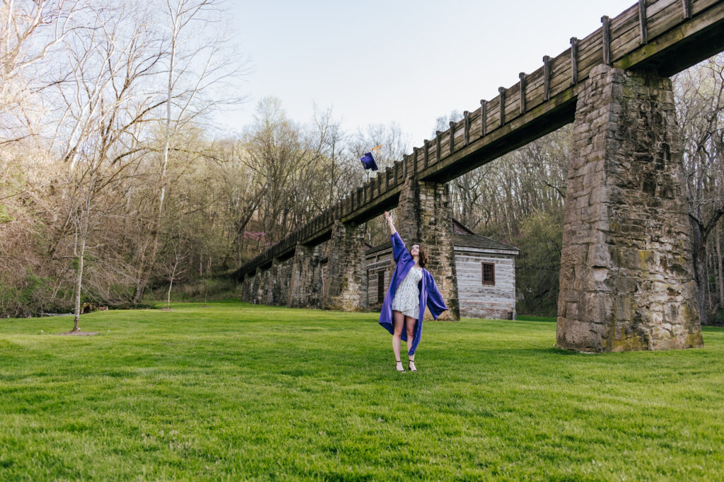 A senior wearing a purple graduation gown tosses her cap in the air while standing in a green lawn in front of a stone bridge
