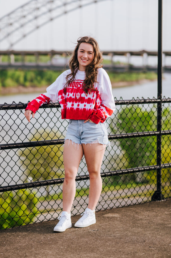 A senior poses in front of the Southern Indiana bridge wearing her college tee and shorts