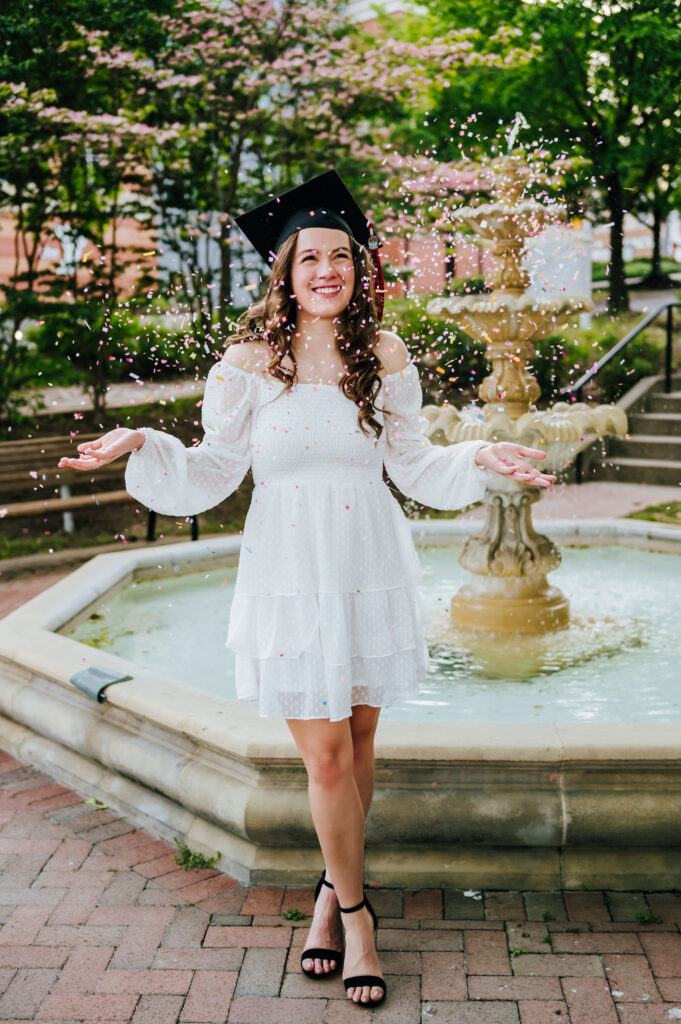 A high school senior throws confetti in the air. She is wearing a white dress and her graduation cap.