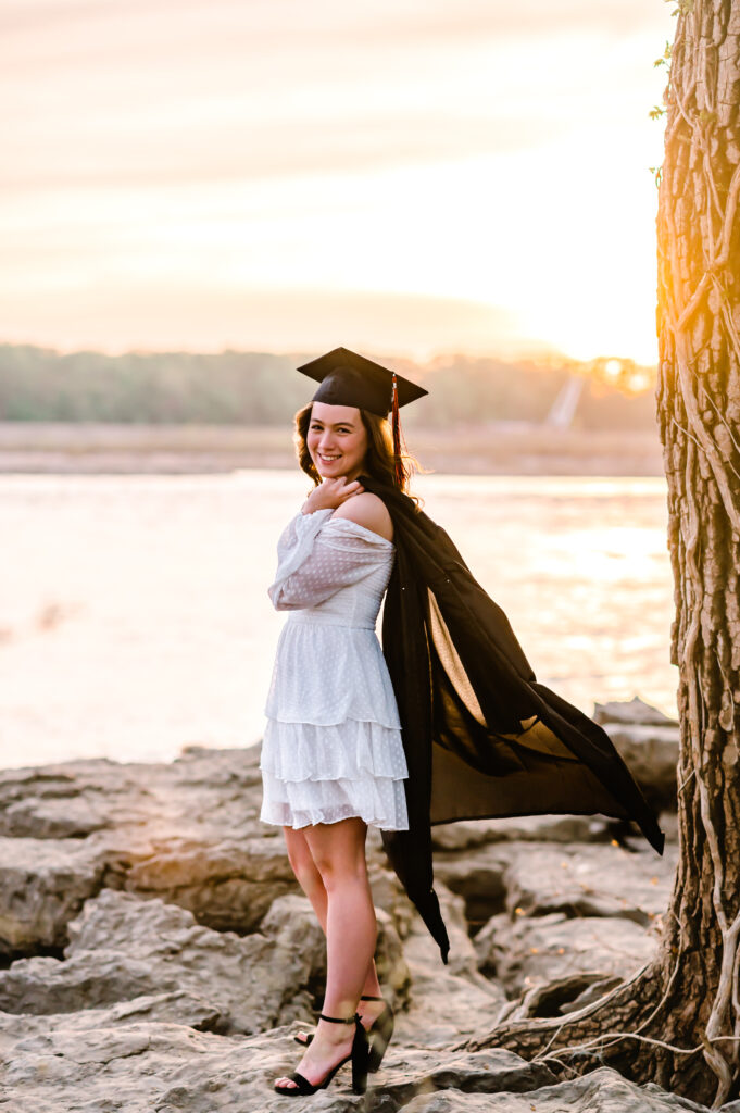 A high school grad in a white dress poses in front of the sunset with her graduation cap on and the robe over her shoulder