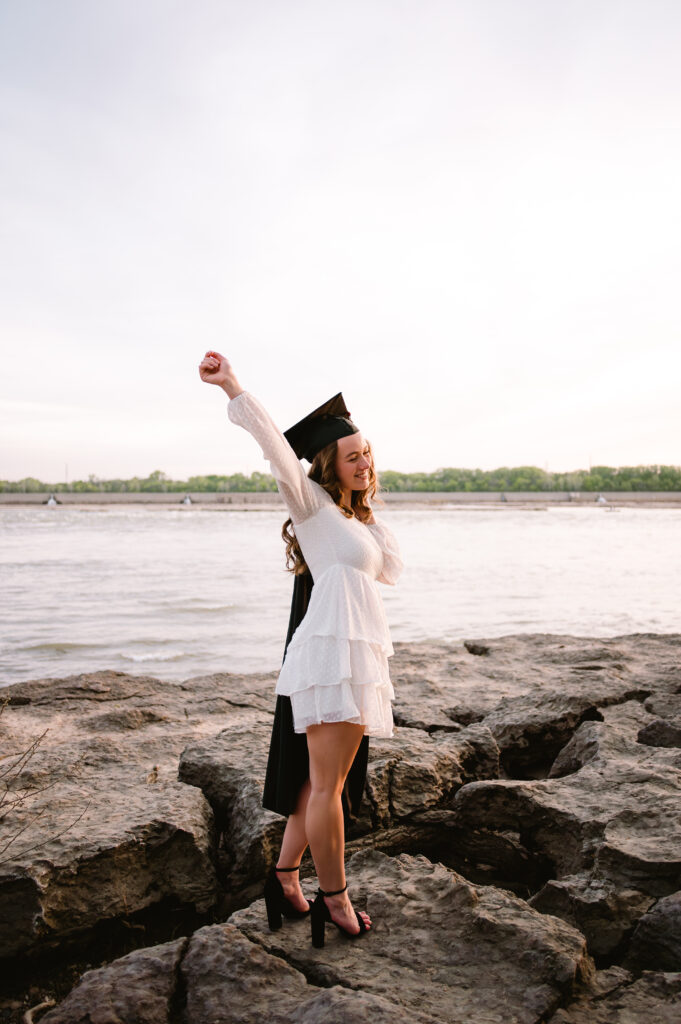 A high school grad in a white dress throws her hands up in celebration with her graduation cap on and the robe over her shoulder