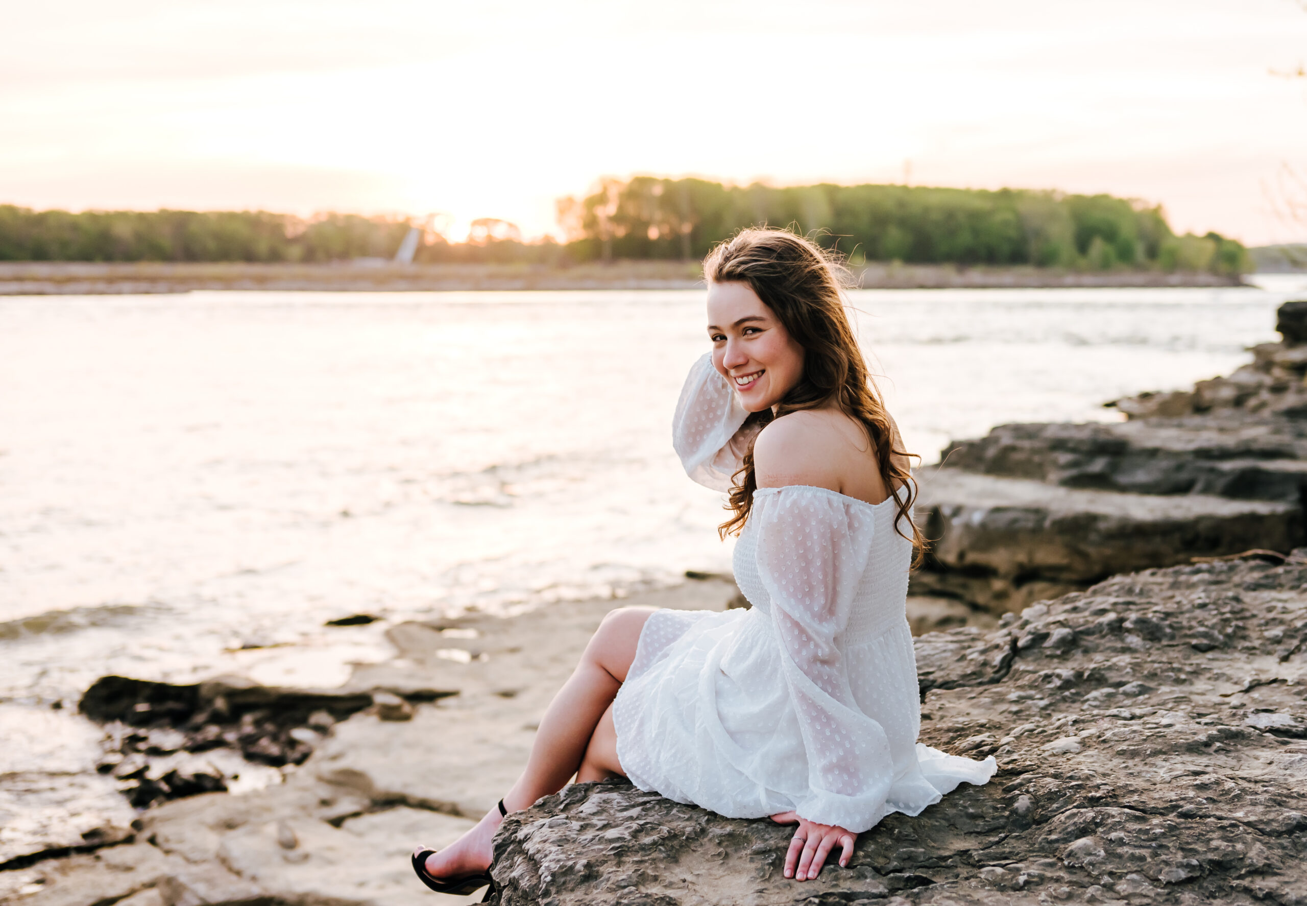 A girl in a white dress smiles, the sun setting behind her, as she sits on a riverside rock overlooking the water.