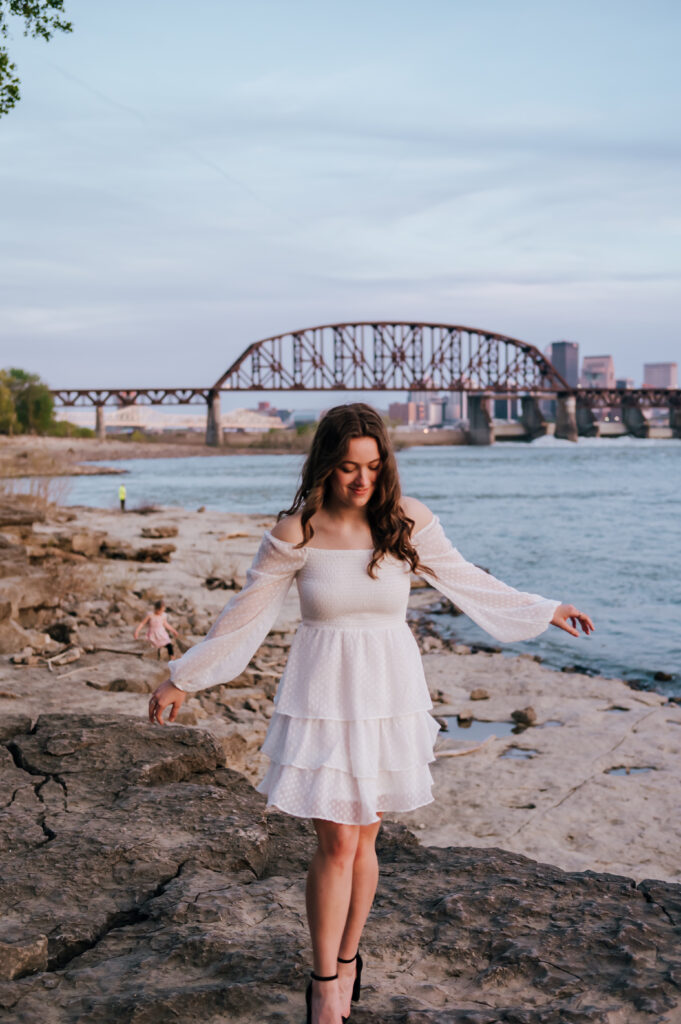 A girl in a white dress walks along the rocks with the Louisville bridge and Ohio River behind her. 