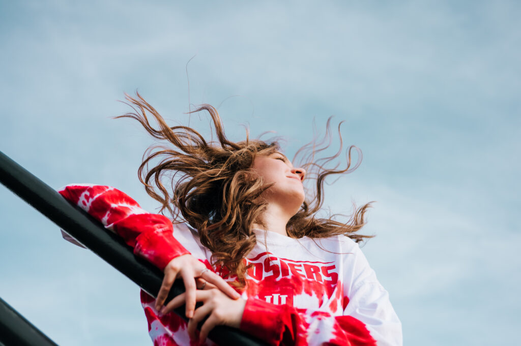 A photo from below of a high school senior with her hair blowing in the wind and the blue sky behind her