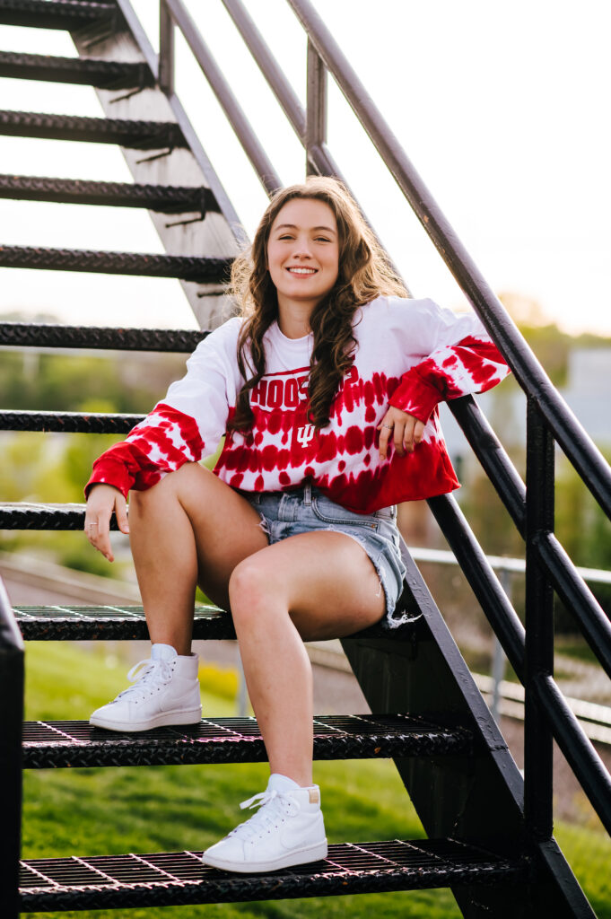 A high school grad wears her college tee and jean shorts. She is seated on a black staircase