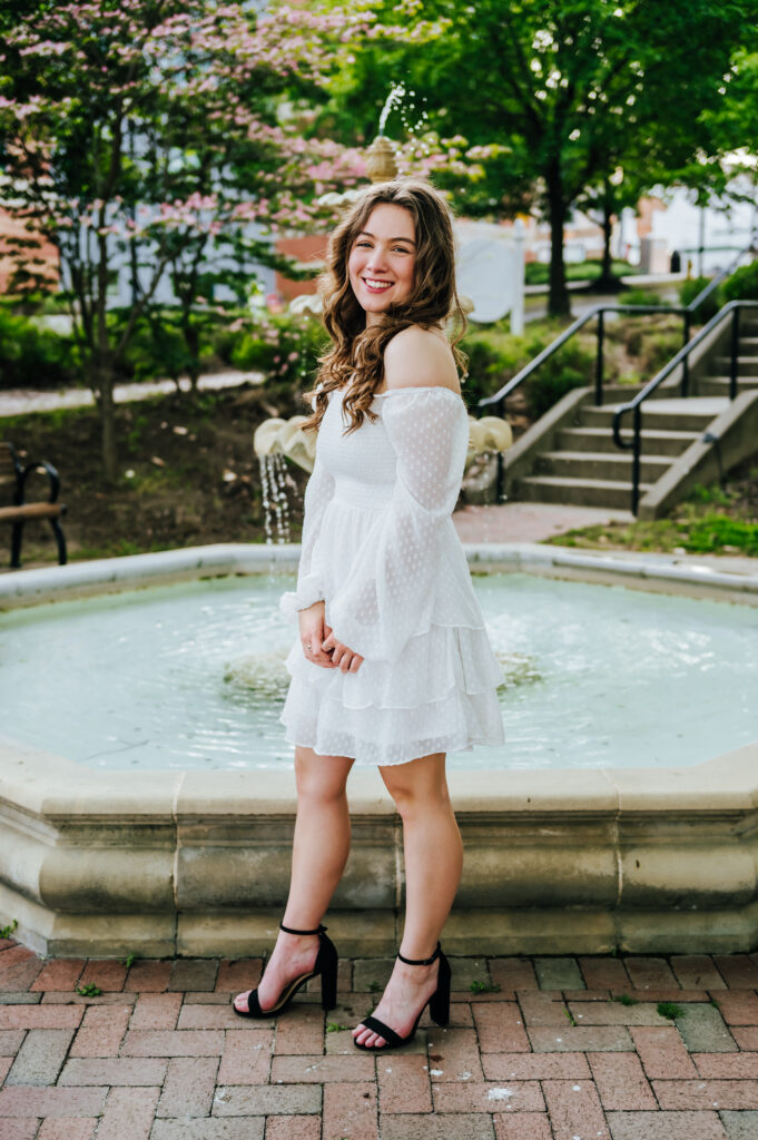 A girl in a white dress clasps her hands together as she stands in front of a fountain
