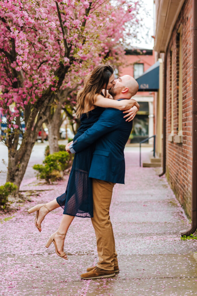 A man picks up his fiancee in front of a pink blossoming tree