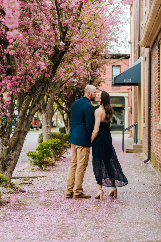 A couple wearing navy blue outfits poses for engagement photos with pink trees in the background