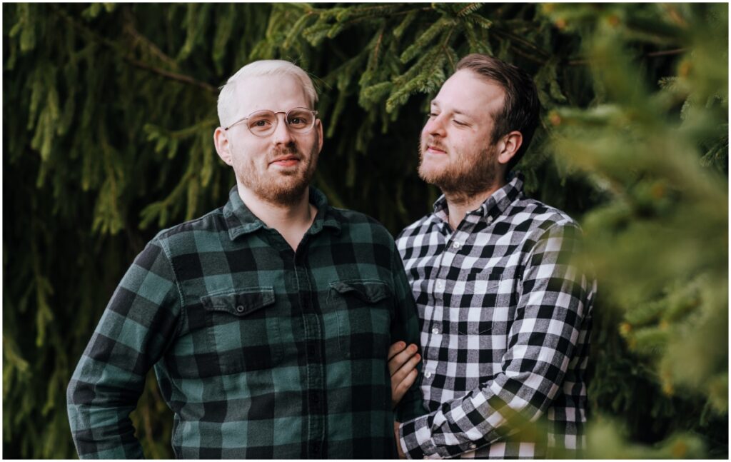 A man looks at his fiance and smiles. Both wear flannel shirts and stand in the midst of evergreen trees.