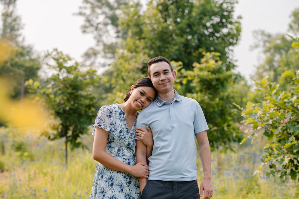 A bride to be rests her head on her fiance's shoulder as they stand in the middle of a beautiful field of tall grass and purple flowers